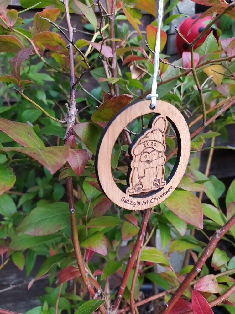 Personalised circular wooden bauble featuring a laser-engraved penguin in the centre, with the year engraved on its hat and "First Christmas" along with a custom name engraved around the edge. Perfect keepsake ornament for the Christmas tree.