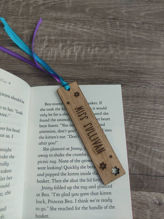 Personalised wooden bookmark engraved with flower designs, featuring two coloured ribbons tied at the top. 