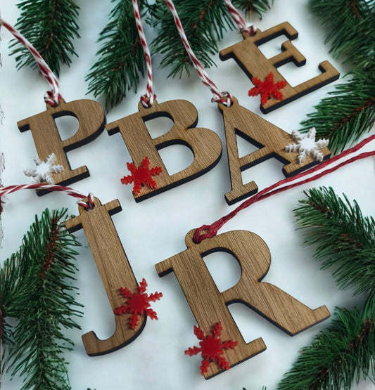 Wooden Christmas tree decorations shaped as letters with red and white snowflake accents, made from oak veneer with festive hanging strings, surrounded by pine branches.