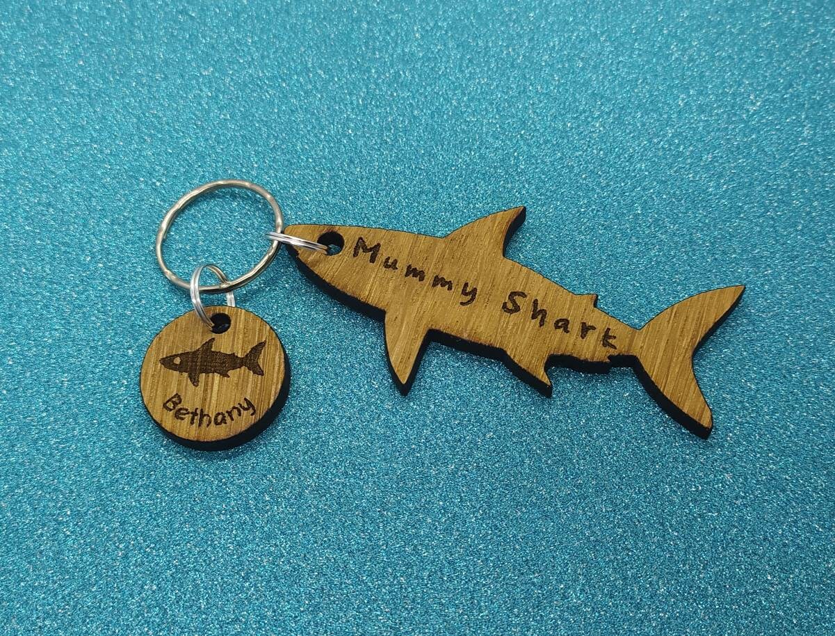 Shark-shaped keyring with "Mummy Shark" engraved, featuring child name charms around the outside and a shark in the middle.