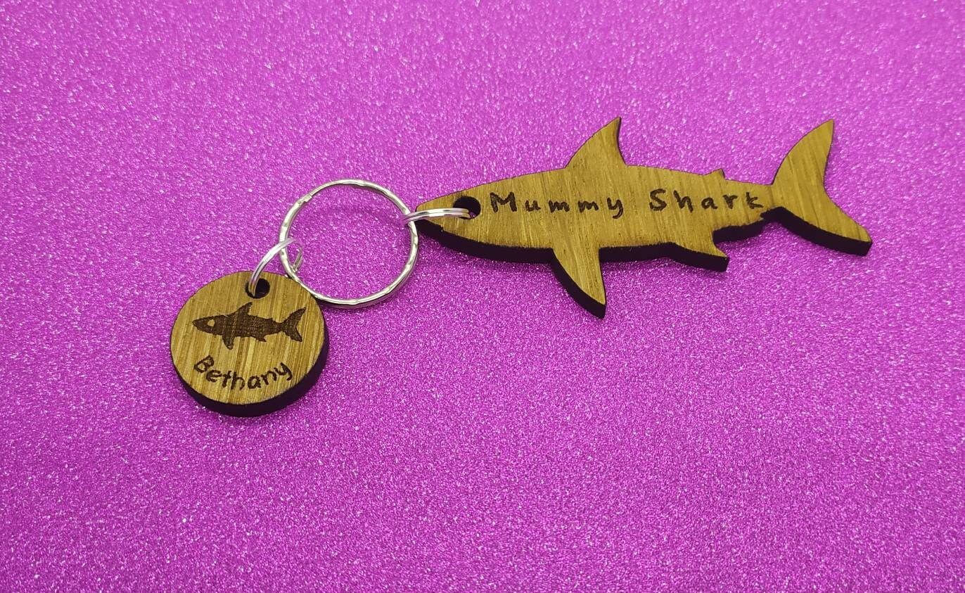 Shark-shaped keyring with "Mummy Shark" engraved, featuring child name charms around the outside and a shark in the middle.