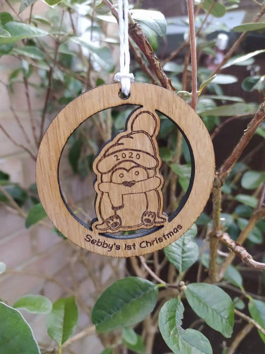 Personalised circular wooden bauble featuring a laser-engraved penguin in the centre, with the year engraved on its hat and "First Christmas" along with a custom name engraved around the edge. Perfect keepsake ornament for the Christmas tree.