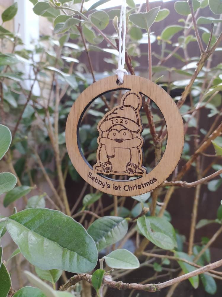 Personalised circular wooden bauble featuring a laser-engraved penguin in the centre, with the year engraved on its hat and "First Christmas" along with a custom name engraved around the edge. Perfect keepsake ornament for the Christmas tree.