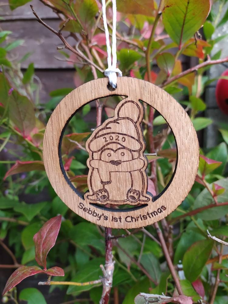 Personalised circular wooden bauble featuring a laser-engraved penguin in the centre, with the year engraved on its hat and "First Christmas" along with a custom name engraved around the edge. Perfect keepsake ornament for the Christmas tree.