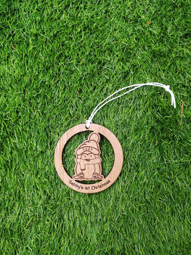 Personalised circular wooden bauble featuring a laser-engraved penguin in the centre, with the year engraved on its hat and "First Christmas" along with a custom name engraved around the edge. Perfect keepsake ornament for the Christmas tree.