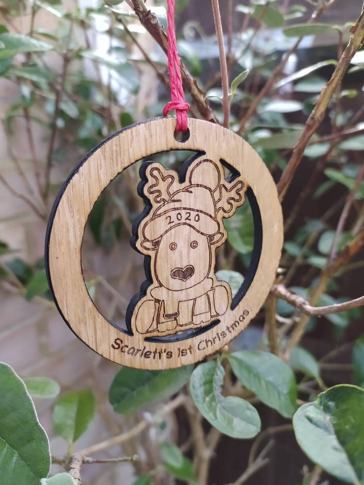 Personalised circular wooden bauble featuring a laser-engraved reindeer in the centre, with the year engraved on its hat and "First Christmas" along with a custom name engraved around the edge. Perfect keepsake ornament for the Christmas tree.