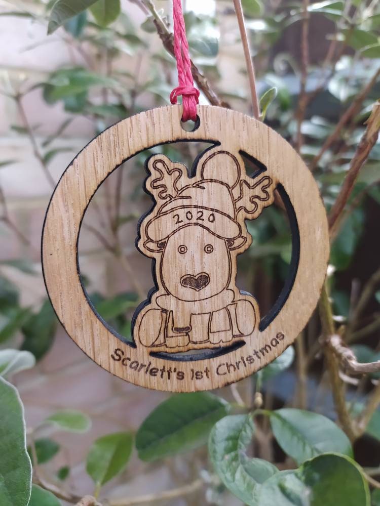 Personalised circular wooden bauble featuring a laser-engraved reindeer in the centre, with the year engraved on its hat and "First Christmas" along with a custom name engraved around the edge. Perfect keepsake ornament for the Christmas tree.