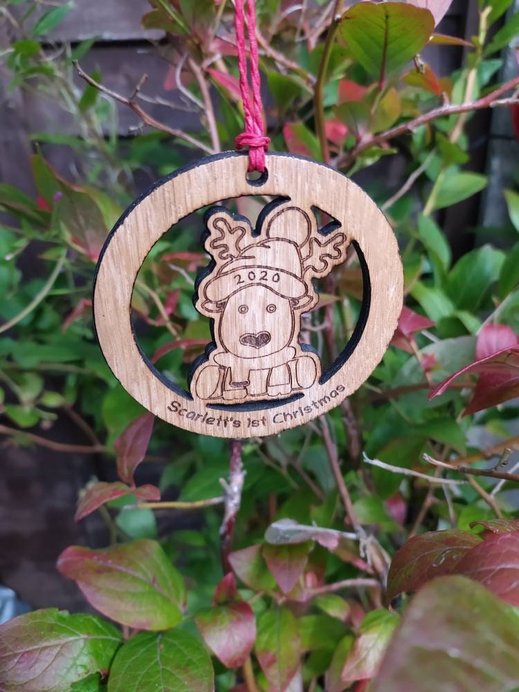 Personalised circular wooden bauble featuring a laser-engraved reindeer in the centre, with the year engraved on its hat and "First Christmas" along with a custom name engraved around the edge. Perfect keepsake ornament for the Christmas tree.