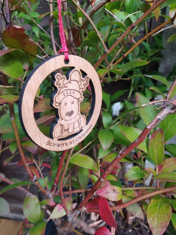 Personalised circular wooden bauble featuring a laser-engraved reindeer in the centre, with the year engraved on its hat and "First Christmas" along with a custom name engraved around the edge. Perfect keepsake ornament for the Christmas tree.